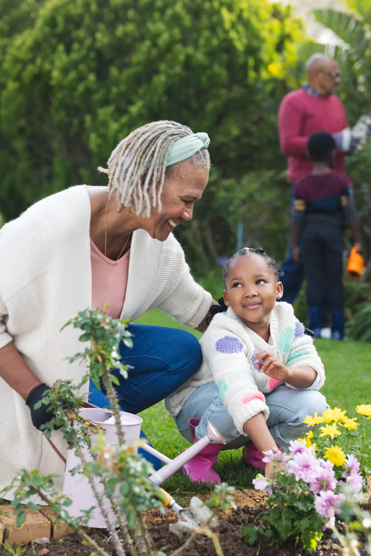 Image of a grandma with their grand daughter in a garden crouching down to water the flowers.