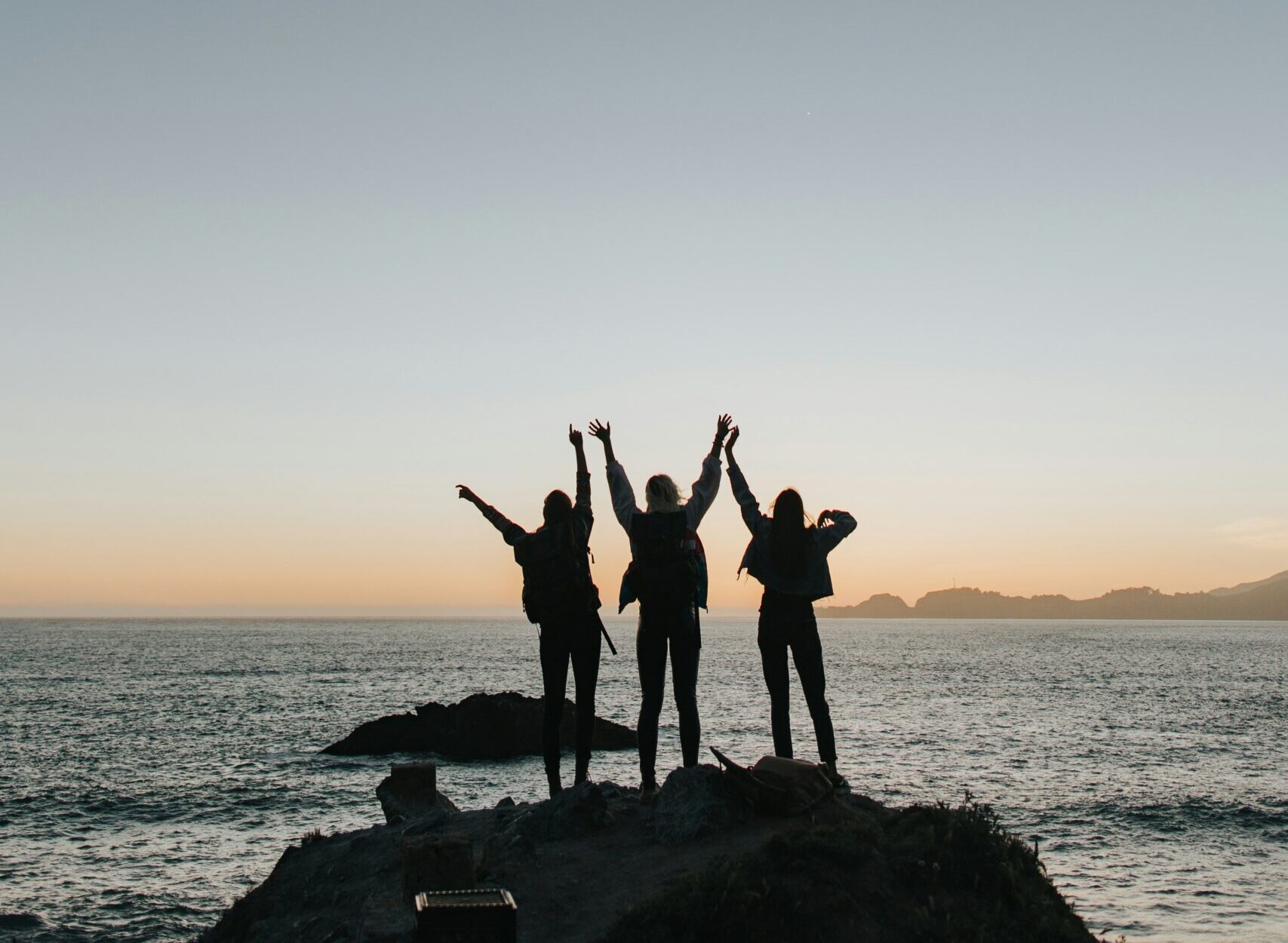 Image of silhouette of three people standing on a rock on shore looking out at the ocean.