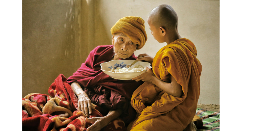 Image of elder Asian woman being fed rice by a young boy in yellow robes.